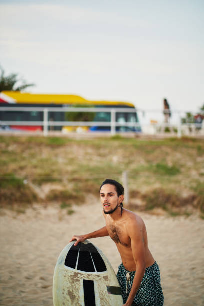 Young athletic man holding his skimboard on the beach, preparing to ride the waves at sunset, with a bus parked in the background, creating a vibrant coastal scene