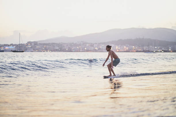 Young man practices skimboarding on the calm sea at sunset, with a city coastline and mountains in the background, enjoying the golden hour light and the thrill of riding the waves