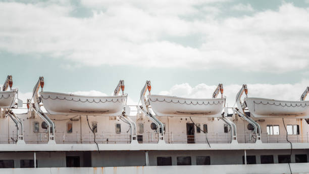 A row of white lifeboats secured on a rusted cruise ship under a partly cloudy sky. The metal davits and pulleys hold the boats in place, while the ship's exterior shows signs of wear