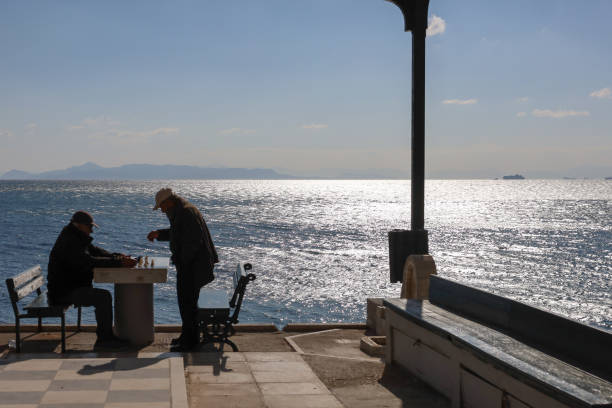 Mature adult man with gray hair and beard sitting by the water, holding a coffee cup, looking thoughtfully at the view, and enjoying a peaceful moment outdoors at a marina