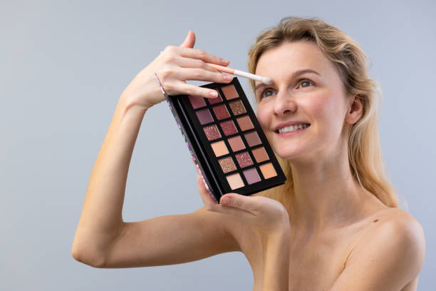 istock A youthful woman applying makeup using a colorful eyeshadow palette while smiling and looking in the mirror, showcasing her beauty routine and self-care practices in a bright setting. 2203306894