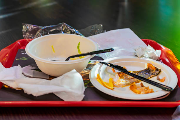Red tray rests on table with empty plates, leftover food, napkins, utensils. Used disposable plates and plastic utensils on red tray in food court aftermath of lunch