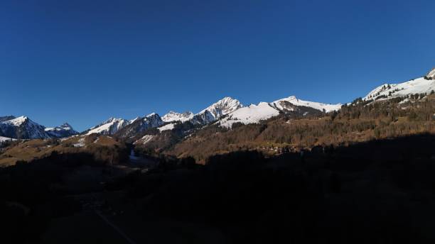 Snow-capped mountains under a clear blue sky, overlooking a valley with a small settlement. Rugged peaks form a dramatic alpine panorama, ideal for nature, travel, and scenic photography enthusiasts .