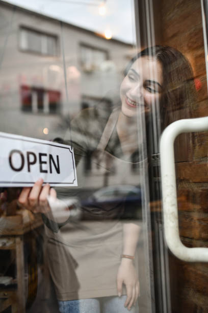 istock Smiling Female Shopkeeper Hanging Open Sign on Storefront Door 2203338009