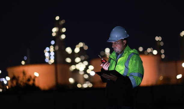 istock Male Middle Eastern engineer wearing a bright green safety jacket, including hard hat examining the survey are using tablet and walkie- talkie to collaborate and discuss screen work plant site use with night lights of oil refinery industry background. 2203352943
