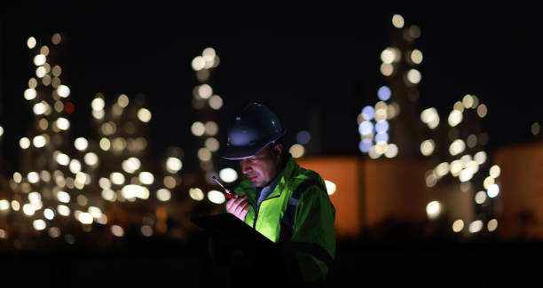 istock Male Middle Eastern engineer wearing a bright green safety jacket, including hard hat examining the survey are using tablet and walkie- talkie to collaborate and discuss screen work plant site use with night lights of oil refinery industry background. 2203352946