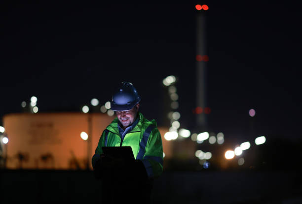istock Male Middle Eastern engineer wearing a bright green safety jacket, including hard hat examining the survey are using tablet and walkie- talkie to collaborate and discuss screen work plant site use with night lights of oil refinery industry background. 2203352950