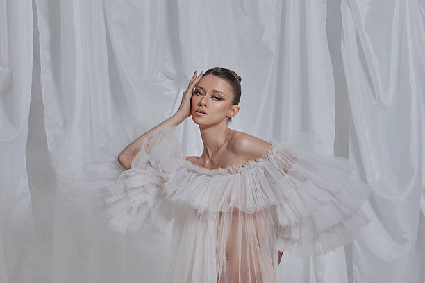 model stands elegantly in an airy white gown featuring ruffled sleeves. backdrop consists of flowing drapery that enhances the delicate ambiance created by soft, diffused lighting