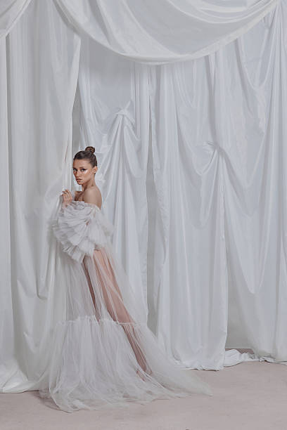 istock model gracefully poses in a flowing white gown adorned with ruffles, standing against softly draped curtains in a bright, airy indoor space, exuding elegance and charm 2203365569