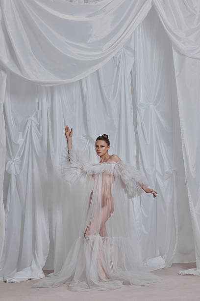 istock model showcases a translucent gown with dramatic sleeves, striking a graceful pose in front of flowing white fabric. setting feels ethereal and artistic, highlighting the garments design 2203365575