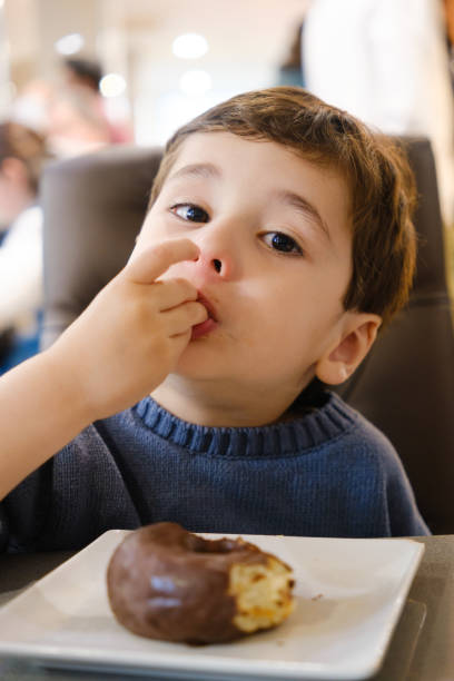 istock Child licking his fingers with a chocolate doughnut in a cafeteria 2203371272