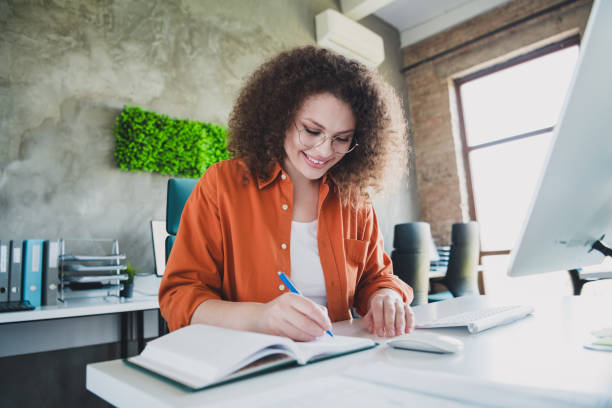 istock Young professional woman working in a modern office, writing notes at a desk while smiling, with natural light and a stylish interior 2203375795