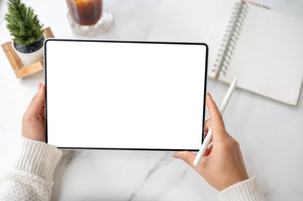 The left hand holding blank screen tablet horizontally with stylus pencil in right hand on the table
