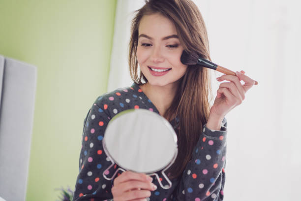 istock Young woman applying makeup while smiling and looking into a handheld mirror in a cozy bedroom, capturing a relaxed morning moment. 2203377505