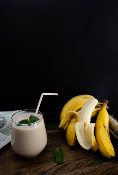 Vertical image of banana smoothie in a glass on a rustic wooden tray next to a bunch of bananas on a dark background. Dark food