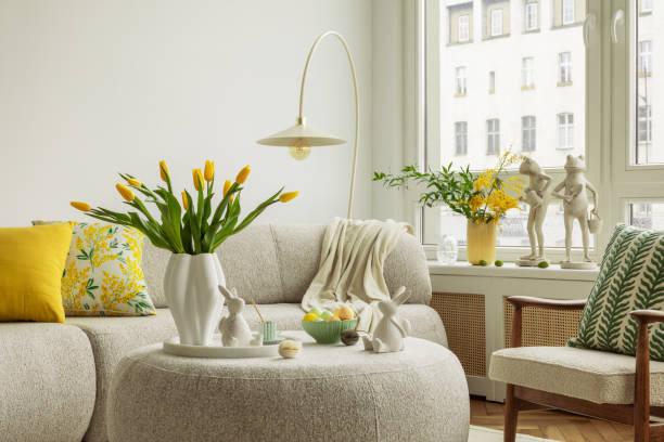 istock Light and airy living space decorated for Easter with soft beige tones, a curved sofa, and a cozy atmosphere. A round ottoman with a marble tray holds pastel eggs, ceramic bunnies, and a cup of coffee. 2203388035
