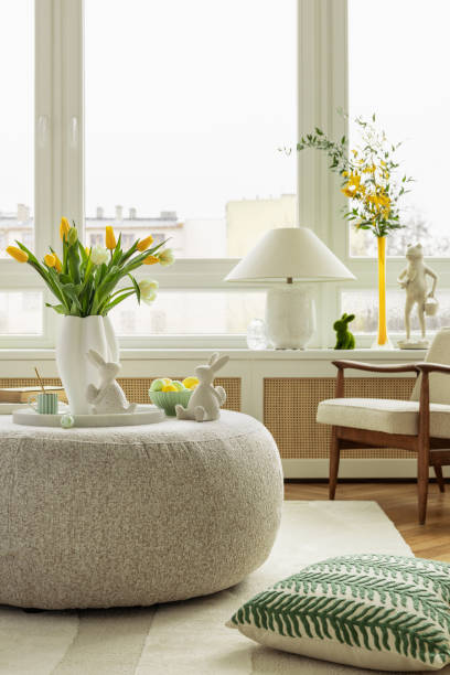istock A modern, minimalist living space with an Easter centerpiece. A round ottoman serves as a display for festive decorations, including ceramic bunnies, pastel eggs, and fresh flowers. 2203388041