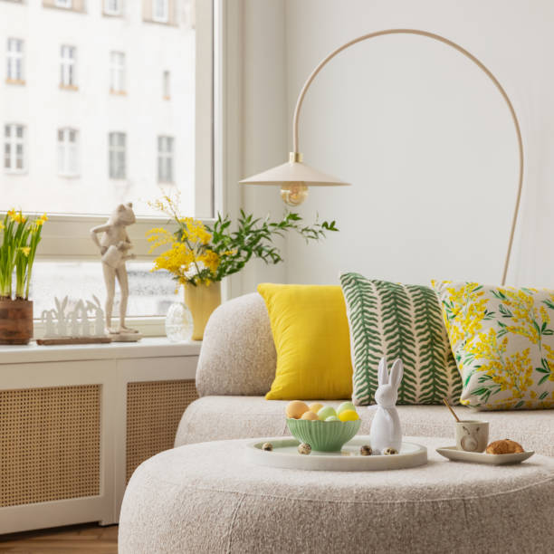 istock Light and airy living space decorated for Easter with soft beige tones, a curved sofa, and a cozy atmosphere. A round ottoman with a marble tray holds pastel eggs, ceramic bunnies, and a cup of coffee. 2203388817
