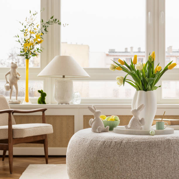 istock Light and airy living space decorated for Easter with soft beige tones, a curved sofa, and a cozy atmosphere. A round ottoman with a marble tray holds pastel eggs, ceramic bunnies, and a cup of coffee. 2203388848