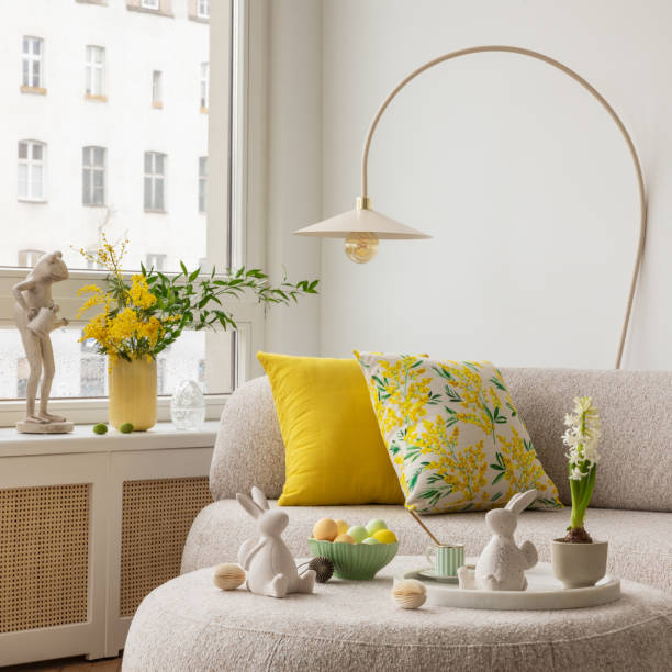 istock Light and airy living space decorated for Easter with soft beige tones, a curved sofa, and a cozy atmosphere. A round ottoman with a marble tray holds pastel eggs, ceramic bunnies, and a cup of coffee. 2203388953