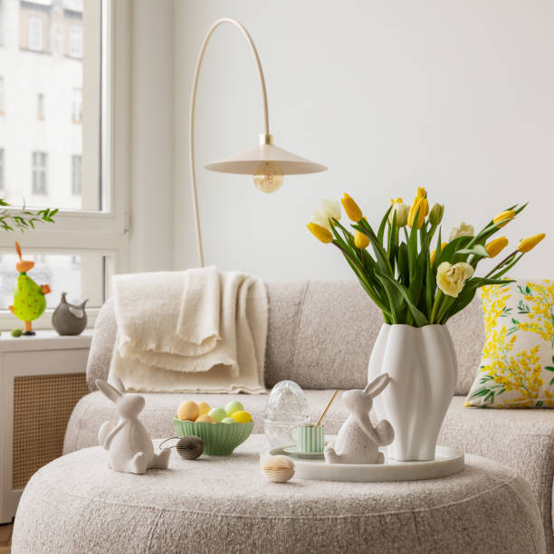 istock Light and airy living space decorated for Easter with soft beige tones, a curved sofa, and a cozy atmosphere. A round ottoman with a marble tray holds pastel eggs, ceramic bunnies, and a cup of coffee. 2203388954