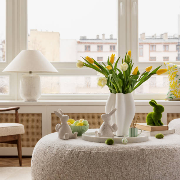 istock Light and airy living space decorated for Easter with soft beige tones, a curved sofa, and a cozy atmosphere. A round ottoman with a marble tray holds pastel eggs, ceramic bunnies, and a cup of coffee. 2203388969