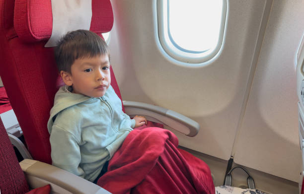 This image captures a moment of contemplation as a young boy sits in an airplane seat, wrapped in a blanket, reflecting on his surroundings while peering out from the window, lost in thought.