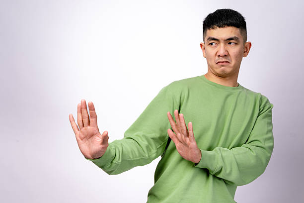 istock An Asian man in a green sweatshirt makes a disgusted expression while holding both hands up in a defensive gesture. He leans slightly backward, looking away. The background is white 2203398227