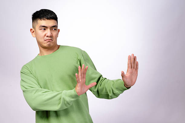 istock An Asian man in a green sweatshirt makes a disgusted expression while holding both hands up in a defensive gesture. He leans slightly backward, looking away. The background is white 2203398684