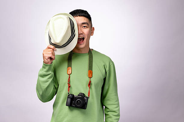 An Asian man in a green sweater playfully holds a fedora hat over one eye while smiling. A camera with a strap hangs around his neck, and he stands against a plain white background