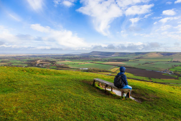 A lone male hiker, wearing raincoat and backpack, looks out over the rolling green landscape of the South Downs in East Sussex, UK, during golden hour. He is sitting on a bench and enjoying the view from the top of a hill.