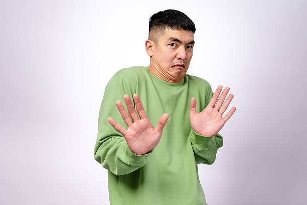 istock An Asian man in a green sweatshirt makes a hesitant expression while raising both hands in a defensive gesture. His eyebrows are raised, and his mouth is slightly pouted. The background is plain white 2203399535