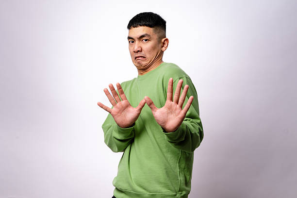 istock An Asian man in a green sweatshirt makes a hesitant expression while raising both hands in a defensive gesture. His eyebrows are raised, and his mouth is slightly pouted. The background is plain white 2203399690