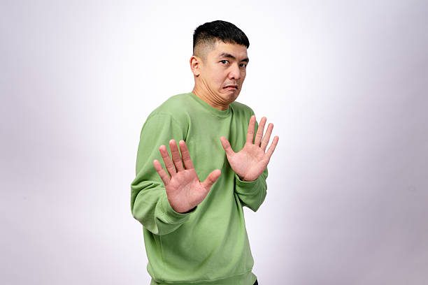 istock An Asian man in a green sweatshirt makes a hesitant expression while raising both hands in a defensive gesture. His eyebrows are raised, and his mouth is slightly pouted. The background is plain white 2203400240