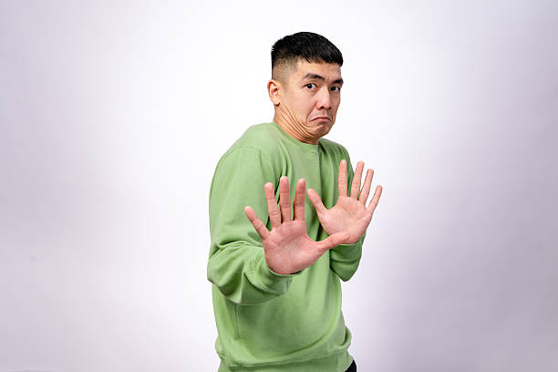istock An Asian man in a green sweatshirt makes a hesitant expression while raising both hands in a defensive gesture. His eyebrows are raised, and his mouth is slightly pouted. The background is plain white 2203400241