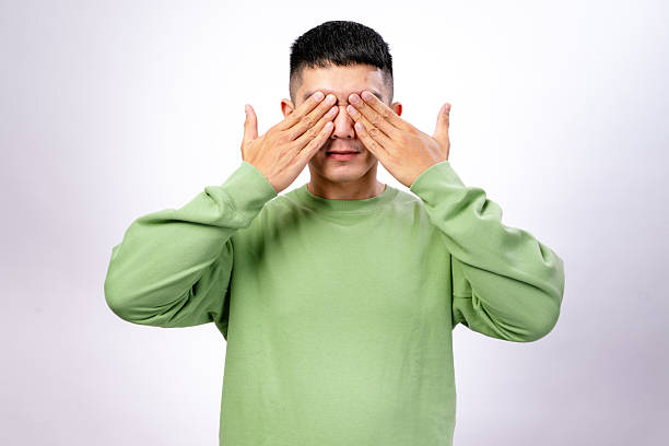 istock An Asian man in a green sweatshirt covers his eyes with both hands while yawning. He appears sleepy or surprised, with his mouth open against a plain white background 2203400682