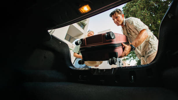 A man loading a suitcase into the open trunk of the car, captured from the inside of the car. Preparing for an upcoming trip with careful packing.
