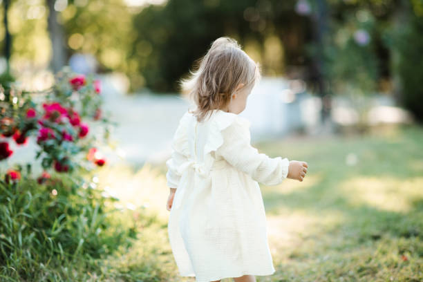 Cute baby girl 1-2 year old wearing white dress walking in rose flowers in garden outdoors rear view. Springtime. Childhood.