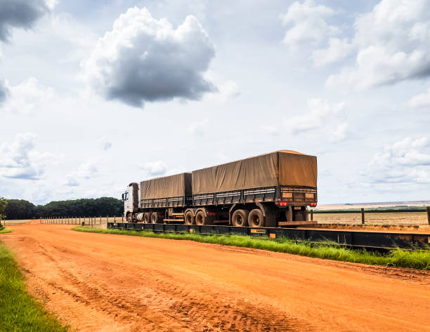 A freight truck with two tarped trailers is positioned on a weighbridge, undergoing weight measurement along a rural dirt road. The scene features open fields and a partly cloudy sky, emphasizing agricultural logistics and rural transport infrastructure.
