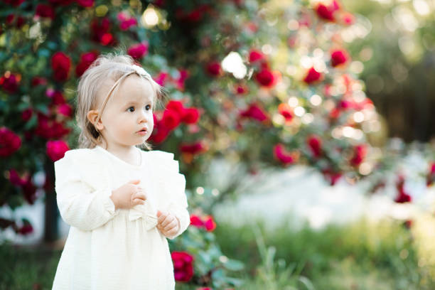 Cute baby girl 1-2 year old wearing white dress posing with rose flowers in garden outdoors. Springtime. Childhood.