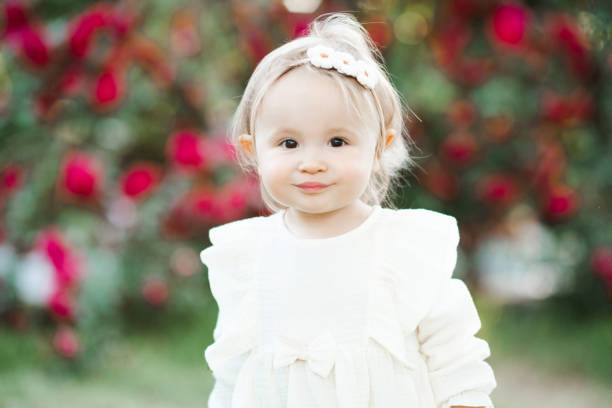 Cute baby girl 1-2 year old wearing white dress posing with rose flowers in garden outdoors. Springtime. Childhood.