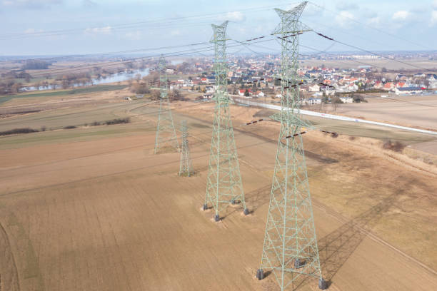 Aerial view of towering electricity pylons stretching across open fields towards a distant village. These structures form a crucial part of the energy infrastructure, seamlessly blending with the rural environment while supplying power to local communities.