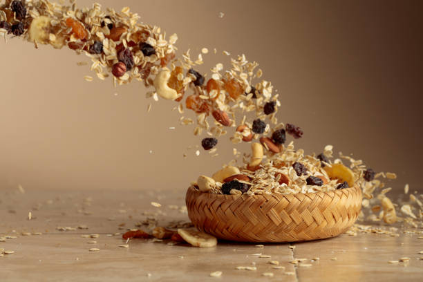 Muesli in motion on a beige background. Oat flakes, assorted nuts, raisins, and dried banana slices fall into a bowl. Copy space.