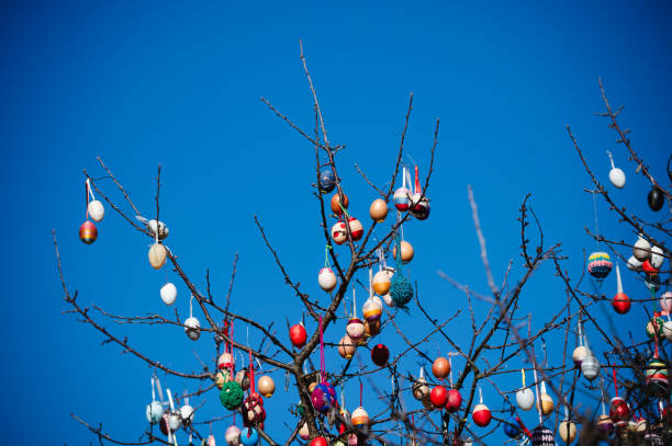 A detailed close-up view of a tree decorated with vibrant Easter eggs hanging from its branches against a bright blue sky. The mix of handmade and painted ornaments adds a festive and cheerful atmosphere, celebrating the joy and tradition of Easter. The intricate details and variety of colors create a visually captivating scene.