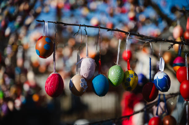 A detailed close-up of brightly colored Easter eggs hanging from a tree branch, illuminated by sunlight. The festive decorations, featuring various shades and patterns, create a joyful and vibrant atmosphere. The blurred background enhances the depth, making the eggs stand out beautifully.