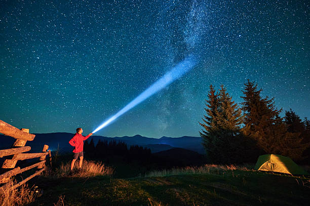 Man hiker stands under dazzling starry sky, shining flashlight into heavens. stock photo