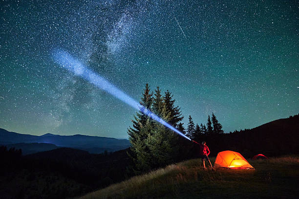 istock Man hiker stands under dazzling starry sky, shining flashlight into heavens. 2203425373