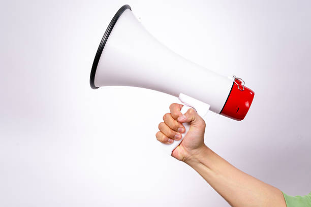 istock A close-up image of a hand holding a white megaphone with a red back against a plain white background. The hand is gripping the handle firmly, and the megaphone is pointed upward. 2203428521