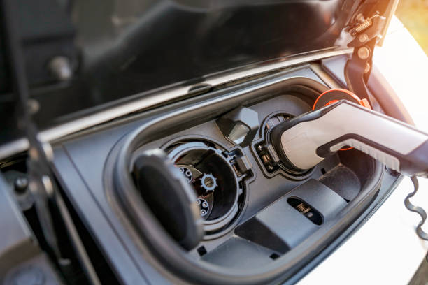 Electric car charging port with two circular sockets is shown. A black and white charging plug is inserted into one of the ports, signaling active charging. The setup is located at the front of the car, with the hood partially open, providing access to the charging area.