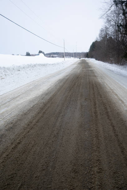 Road covered with muddy frozen slush in a snowy landscape with telephone poles and a barn in the distance. Photographed in Aroostook County,  Maine.